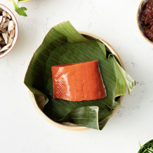 A bamboo steamer basket lined with a banana leaf and an orange uncooked sockeye salmon portion positioned in the center.
