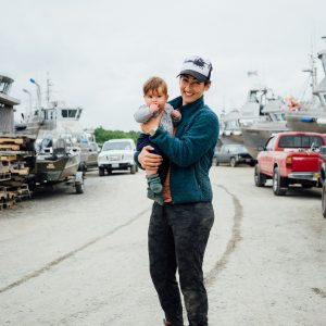 A woman holding a baby in the boatyard, boats and trucks in the background.