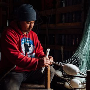 A fisherman sitting on a wooden bench repairing a net with a weaving shuttle in his hand.