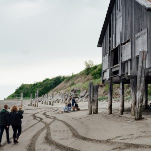A couple with a baby walking on a beach toward a group of people sitting around a fire, tire tracks visible in the sand and an old wooden structure off to the right.