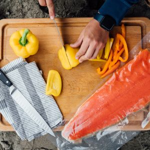 A bird's eye view of a person slicing bell peppers on a wooden cutting board resting in the sand, a packaged sockeye salmon fillet on the right and a fillet knife and cloth napkin on the left.