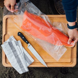 A bird's eye view of a person removing a sockeye salmon fillet from a vacuum sealed back on a wooden cutting board resting on the sand, a fillet knife and cloth napkin on the side.