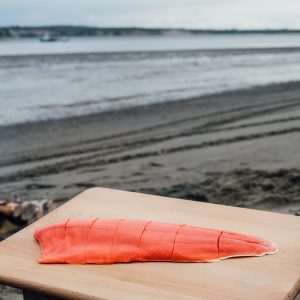 A sockeye salmon fillet cut into portions on a wooden cutting board resting on a beach with the water in the background.