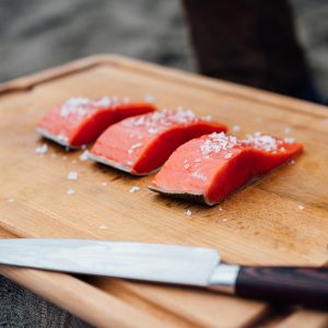 A wooden cutting board with three bright uncooked sockeye salmon portions sprinkled with gourmet sea salt.