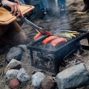 A beach campfire with a metal grill grate on top and a man placing a sockeye salmon fillet on the grill with tongs, a wooden cutting board in his lap.