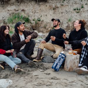 A group of people sitting in a semicircle on a beach enjoying jarred salmon on crackers, all laughing and smiling.
