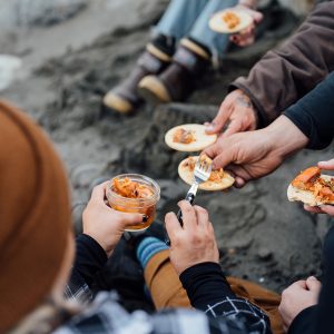 A group of people sitting on a beach serving jarred salmon onto crackers.