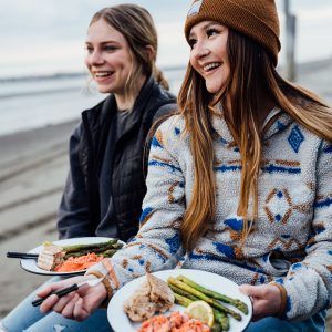 Two women sitting on a beach with plates of salmon and veggies in their laps, both smiling and laughing.