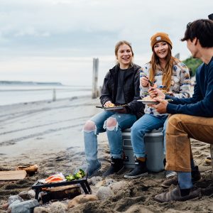 People sitting on a cooler and wood on a beach around a bonfire with plates of salmon in their hands.