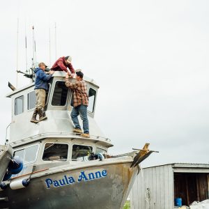 Three fishermen making adjustments on the F/V Paula Anne in a boatyard.