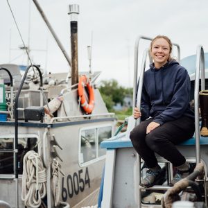 A woman sitting on a boat in a boatyard wearing a navy hoodie, black pants and sandals.