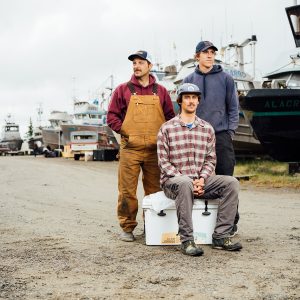 Three fishermen standing and sitting in front of several boats and vehicles in a boatyard.