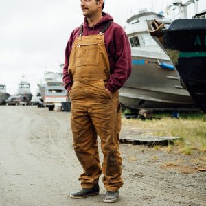 A fisherman wearing brown Carhartt overalls and a maroon hoodie standing in front of several boats in a boatyard with his hands in his pockets.