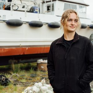 A woman wearing a black jacket standing in front of a boat in a boatyard.