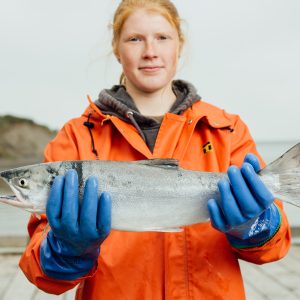 A female fisherman holding a whole sockeye salmon while standing on a dock wearing an orange rain jacket and blue rubber gloves.