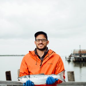 A fisherman holding a whole sockeye salmon while standing on a dock wearing an orange rain jacket and blue rubber gloves.