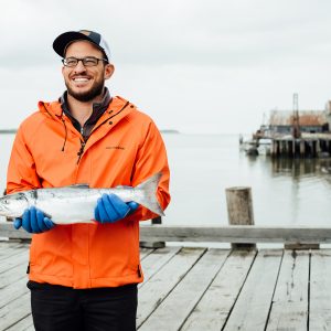A fisherman holding a whole sockeye salmon while standing on a dock wearing an orange rain jacket and blue rubber gloves.