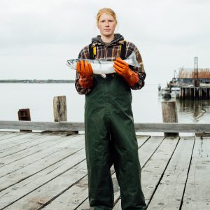 A female fisherman holding a whole sockeye salmon while standing on a dock wearing green waterproof overalls and orange rubber gloves.