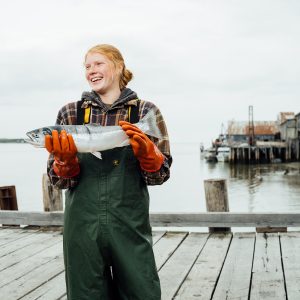 A female fisherman holding a whole sockeye salmon while standing on a dock wearing green waterproof overalls and orange rubber gloves.