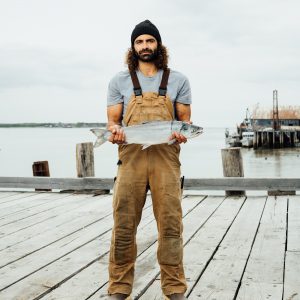 A fisherman holding a whole sockeye salmon while standing on a dock wearing Carhartt overalls and a black beanie.