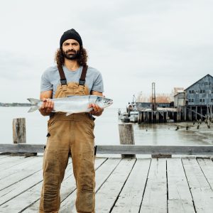 A fisherman holding a whole sockeye salmon while standing on a dock wearing Carhartt overalls and a black beanie.