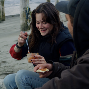 A woman sitting on a beach eating salmon out of a jar while a man in the foreground eats salmon on a cracker.