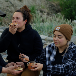 Two women eating jarred salmon on a beach.