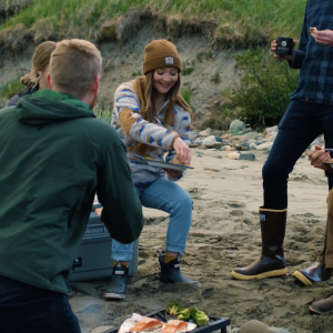 People standing and sitting on a beach with plates of salmon, laughing and enjoying themselves.
