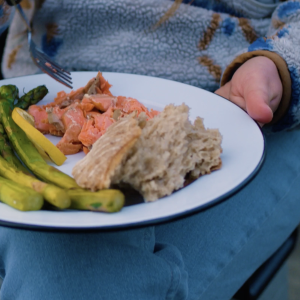 A white plate resting on a woman's lap loaded with veggies, crusty bread and sockeye salmon.