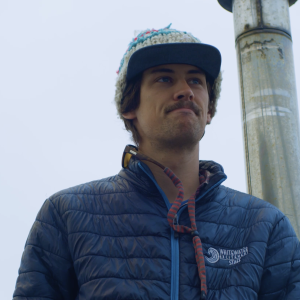 A mustached man wearing a blue puffer jacket and knitted hat with a brim.