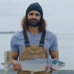 A fisherman on a dock wearing canvas overalls, a blue t-shirt and a navy beanie holding a whole sockeye salmon.