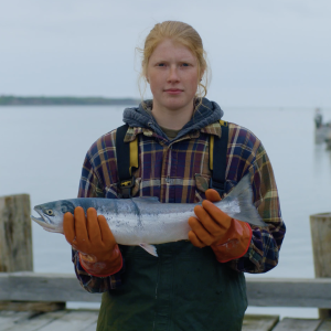 Female fisherman standing on a dock wearing a plaid jacket and orange rubber gloves holding a whole sockeye salmon.