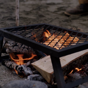 A beach camp fire with a metal cooking grate on top, flames flickering into the grate holes.