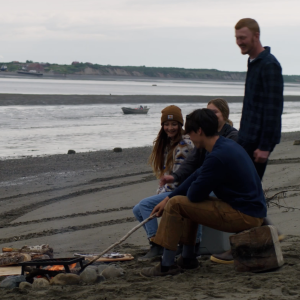Group of people sitting and standing on a beach beside a camp fire, one poking the fire with a stick.