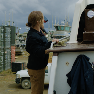 A woman holding on to the side of a boat with a power drill in her left hand.