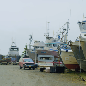 Several boats in a boatyard with trucks and vans parked next to them.