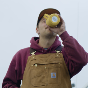 A man drinking from a yellow mug wearing brown Carhartts and a maroon hoodie.