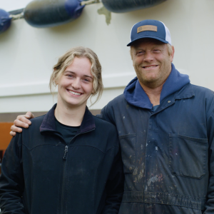 Female and male fishermen posing in a shipyard, the man with his arm around the woman.