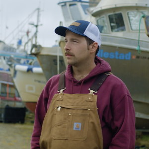 Fisherman standing in a boatyard wearing Carhartt overalls and a maroon hoodie.
