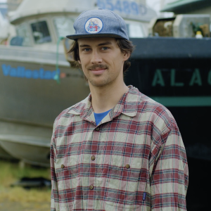 Fisherman wearing a plaid shirt and a blue cap standing in front of boats in the shipyard.