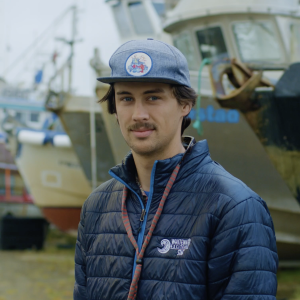 Fisherman wearing a navy down jacket and blue cap standing in front of several boats in the shipyard.