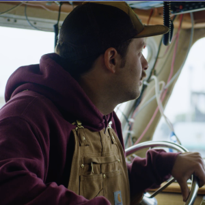 A fisherman in Carhartt overalls and a maroon hoodie wearing a cap sitting behind the steering wheel of a boat and looking out the front window.